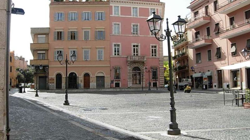 Piazza urbana pavimentata in sampietrini con lampioni in ferro battuto, circondata da palazzi a più piani color rosa con persiane e balconi. Al centro si nota un edificio con ingresso ad arco e finestre regolari, affiancato da altri edifici residenziali con negozi al piano terra.