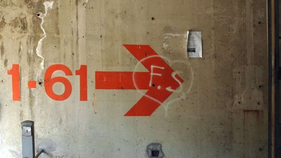 Parete interna in cemento grezzo e macchiato, con una grande freccia rossa dipinta e la scritta “1–61” che indica una direzione. Sulla parete sono presenti segni di usura, un piccolo avviso affisso e alcune prese o elementi metallici incassati.