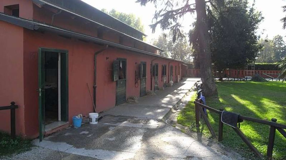 Edificio rurale a un piano con facciata rosa e una fila di porte e finestre lungo un vialetto in cemento. Accanto si vede un prato recintato con alberi alti e una struttura recintata sullo sfondo.