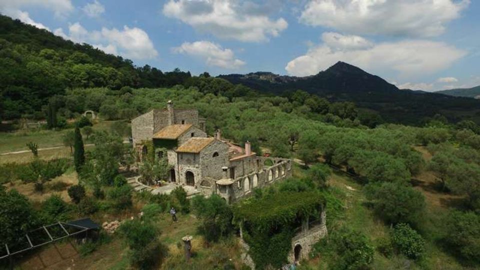 Veduta aerea di un casale in pietra con tetti in tegole e un’area porticata ad archi, circondato da uliveti e vegetazione collinare. Sullo sfondo si vedono montagne boscose sotto un cielo parzialmente nuvoloso.