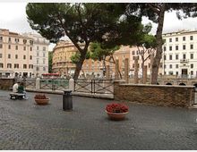 Piazza urbana lastricata con sampietrini, alberi ad alto fusto e fioriere circolari; lungo il bordo ci sono una ringhiera metallica e un muretto in mattoni. Sullo sfondo si vedono palazzi storici di più piani affacciati sulla piazza.