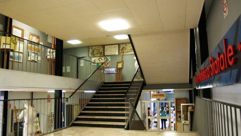 Atrio interno di un edificio pubblico con pavimento in marmo e un’ampia scalinata centrale con corrimano in metallo che conduce al piano superiore. Lungo le pareti e sui ballatoi sono visibili pannelli espositivi e una scritta a parete, con luci a soffitto e accesso a una sala sul fondo.