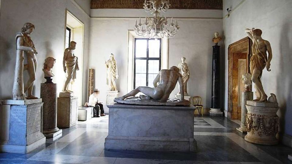 Sala museale con pavimento in marmo a riquadri e soffitto decorato con lampadari, con un grande gruppo scultoreo su piedistallo al centro. Lungo le pareti sono disposte statue classiche su basamenti, con una grande finestra sul fondo e una porta monumentale laterale.