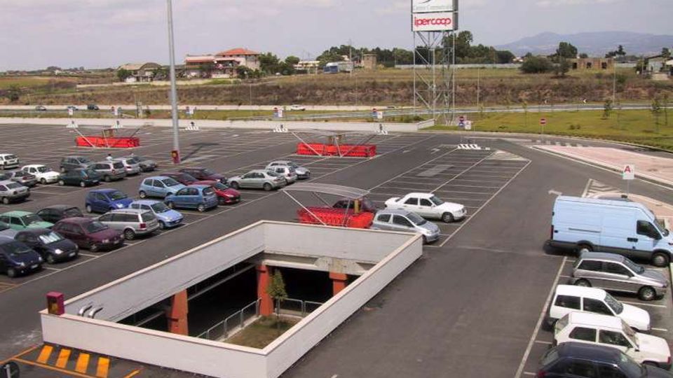 Ampio parcheggio asfaltato di un centro commerciale, con posti auto segnati, diverse vetture e aree con carrelli rossi; sullo sfondo si vedono un’insegna alta e una strada con campagna e colline lontane.