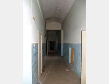 Corridoio interno lungo e stretto con soffitto a volta, pareti bianche e rivestimento di piastrelle azzurre nella parte inferiore. Porte laterali lungo il corridoio e pavimento scuro, con segni di usura e illuminazione fioca verso il fondo.