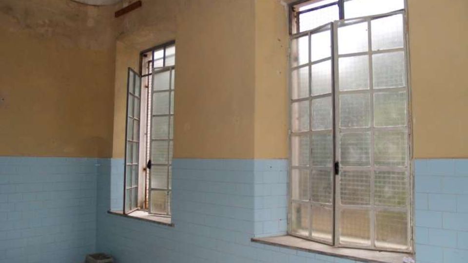 Interno di una stanza spoglia con pareti bicolore (giallo sopra e piastrelle azzurre sotto) e due grandi finestre a riquadri con vetro opaco, una delle quali aperta. Il soffitto è alto e le finestre danno su un ambiente esterno indistinto.