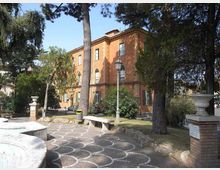 Edificio storico a tre piani con facciata color terracotta e finestre ad arco al piano terra, immerso in un giardino con alti alberi. In primo piano si vede un vialetto pavimentato con motivo a semicerchi, una panchina in pietra e un lampione, con muretti e vasi decorativi ai lati.