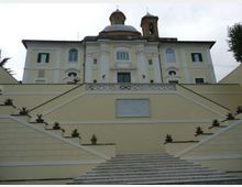 Facciata di un edificio religioso in stile classico con cupola e campanile, vista dal basso. In primo piano una lunga scalinata in pietra con balaustre e terrazze laterali decorate da vasi di piante.