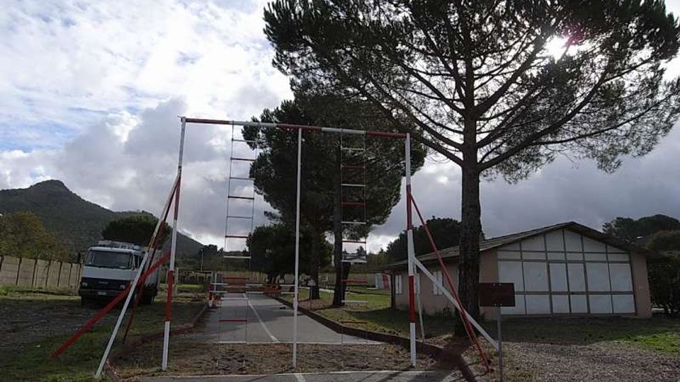 Strada asfaltata in un’area aperta con un telaio metallico bianco e rosso sopra la carreggiata, simile a un ostacolo o percorso di addestramento. Ai lati ci sono alti pini, un edificio basso con pannelli chiari e un camion parcheggiato, con colline sullo sfondo sotto un cielo nuvoloso.