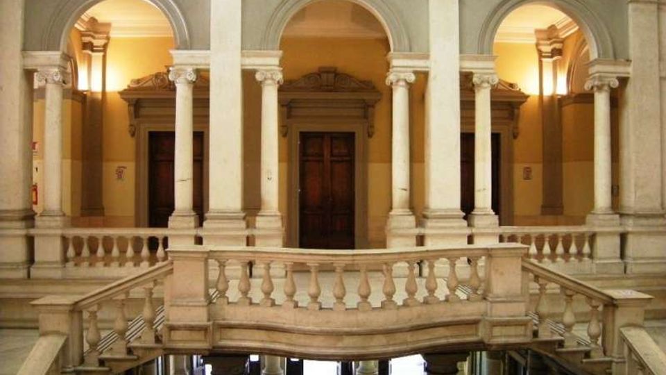 Interno di un edificio storico con loggiato a due livelli, archi a tutto sesto e colonne in pietra. Al centro una balconata con balaustra e scale laterali conduce a un corridoio con porte in legno illuminato da luci calde.
