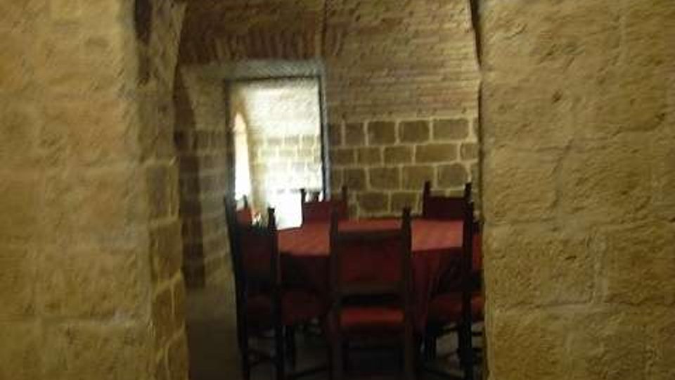 Passaggio ad arco in pietra con soffitto a volta in mattoni, che conduce a una sala con pareti in blocchi di pietra. All’interno si vedono tavoli con tovaglie rosse e sedie in legno, con luce naturale proveniente da una finestra sul fondo.
