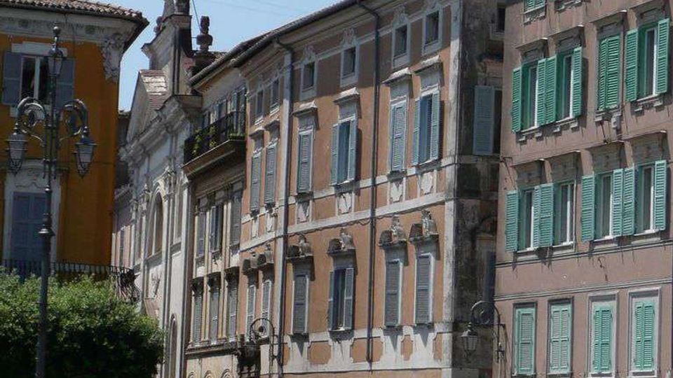 Strada urbana in un centro storico con edifici a più piani dalle facciate intonacate e persiane verdi, finestre regolari e dettagli decorativi. Sul lato della strada si vedono auto parcheggiate, lampioni, vasi con piante e in fondo un campanile dietro i tetti in tegole.