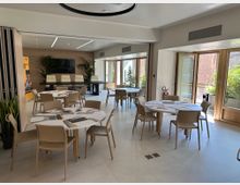 Sala riunioni moderna e luminosa con tavoli rotondi e sedie beige su pavimento chiaro, parete in legno con schermo TV e piante decorative. Grandi porte-finestre con infissi in legno si aprono su una terrazza con verde esterno.