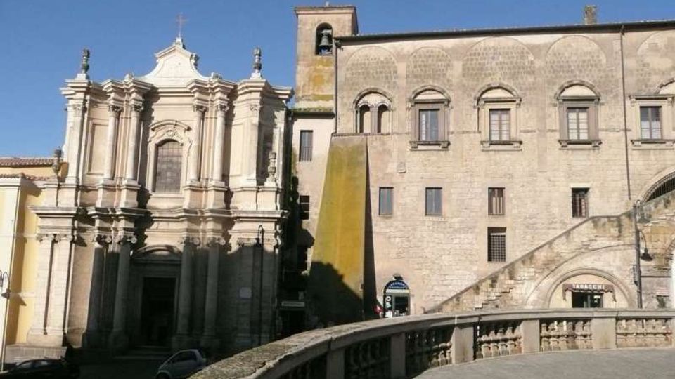 Facciata di una chiesa barocca in pietra chiara con colonne e timpano, accanto a un grande edificio storico in muratura con finestre ad arco e campanile. In primo piano si vede una terrazza o ponte in pietra con balaustra curva e una rampa di scale sul lato destro, sotto cielo sereno.