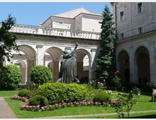 Cortile interno con giardino curato, aiuole fiorite e siepi, circondato da portici ad archi in pietra e balaustra superiore. Al centro si trova una grande statua in bronzo, con edifici in muratura e un alto albero sempreverde sullo sfondo.