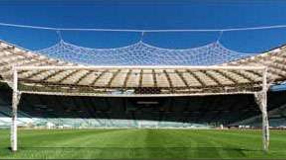 Interno di uno stadio all’aperto visto dal campo, con ampia area di prato verde e tribune semicircolari sullo sfondo. Copertura leggera con struttura reticolare e pilastri bianchi che sorreggono una rete sopra la zona centrale.
