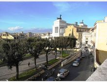 Veduta urbana con una strada in salita costeggiata da auto parcheggiate e alberi, affiancata da una terrazza con muretti e sculture. Sullo sfondo si distingue un grande edificio monumentale bianco con colonne e quadrighe in cima, circondato da palazzi storici.