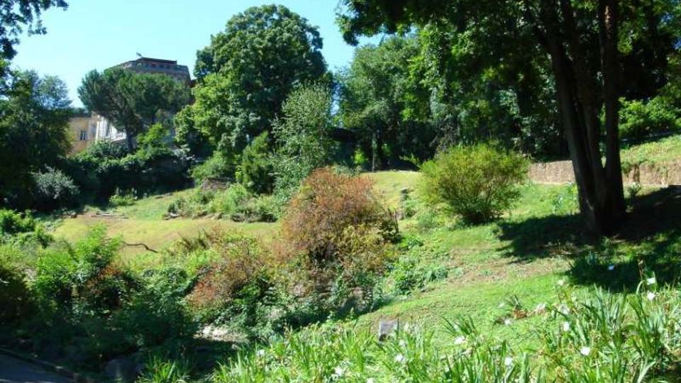 Parco in pendenza con prati, cespugli e aiuole fiorite, circondato da alberi ad alto fusto che creano zone d’ombra. Sullo sfondo si intravedono edifici e un basso muro di contenimento lungo il versante.