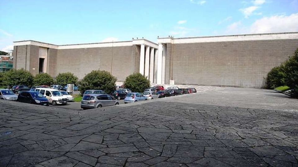 Grande edificio moderno in cemento con facciata lunga e minimale, interrotta da un portale e da alte colonne verticali. In primo piano si vede una piazza lastricata in pietra con parcheggio di auto e alberi ai margini sotto un cielo parzialmente nuvoloso.