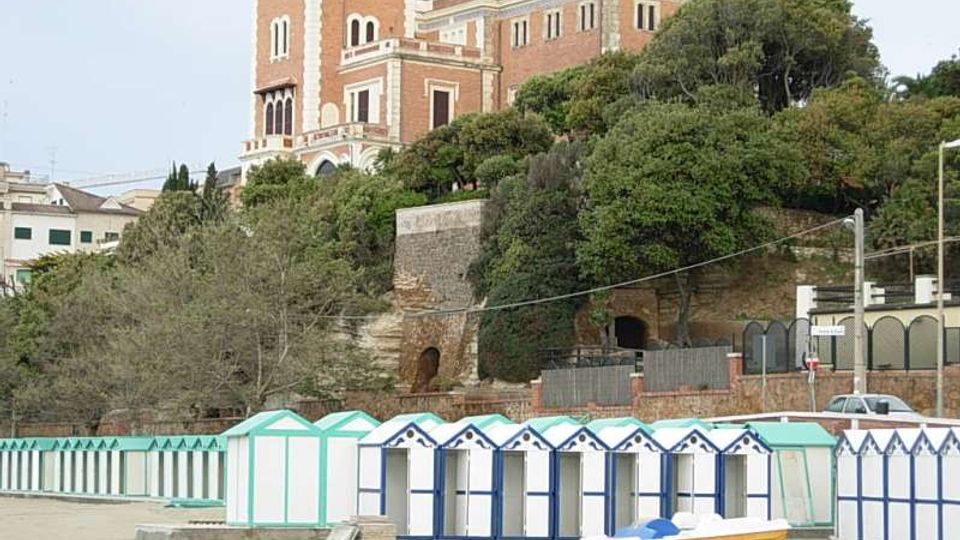 Edificio storico in mattoni con torre quadrata e finestre ad arco, situato su una collina alberata sopra un muraglione. In primo piano si vede una spiaggia con file di cabine balneari bianche e azzurre e una piccola barca sulla sabbia.
