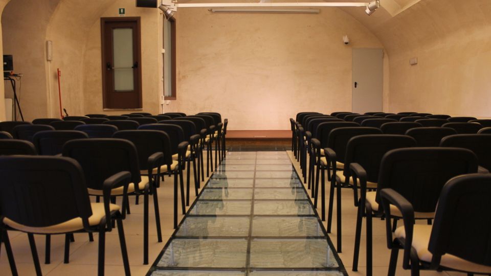 Sala conferenze con soffitto a volta e file di sedie nere disposte ai lati di un corridoio centrale con pavimentazione in vetro. In fondo si vedono una parete chiara, una porta e illuminazione a binario sul soffitto.