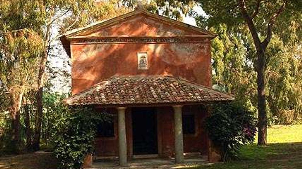 Piccola chiesa rurale in muratura intonacata color rosso, con facciata a timpano e portico d’ingresso con due colonne e tetto in coppi. L’edificio è immerso in un’area verde alberata con prato e alti pini tutt’intorno.