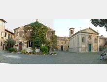 Piazza acciottolata in un borgo storico, circondata da edifici in pietra e intonaco. A destra si vede una chiesa con facciata classica, rosone e campanile, mentre a sinistra c’è una casa rustica coperta di edera con vasi di piante lungo il muro.