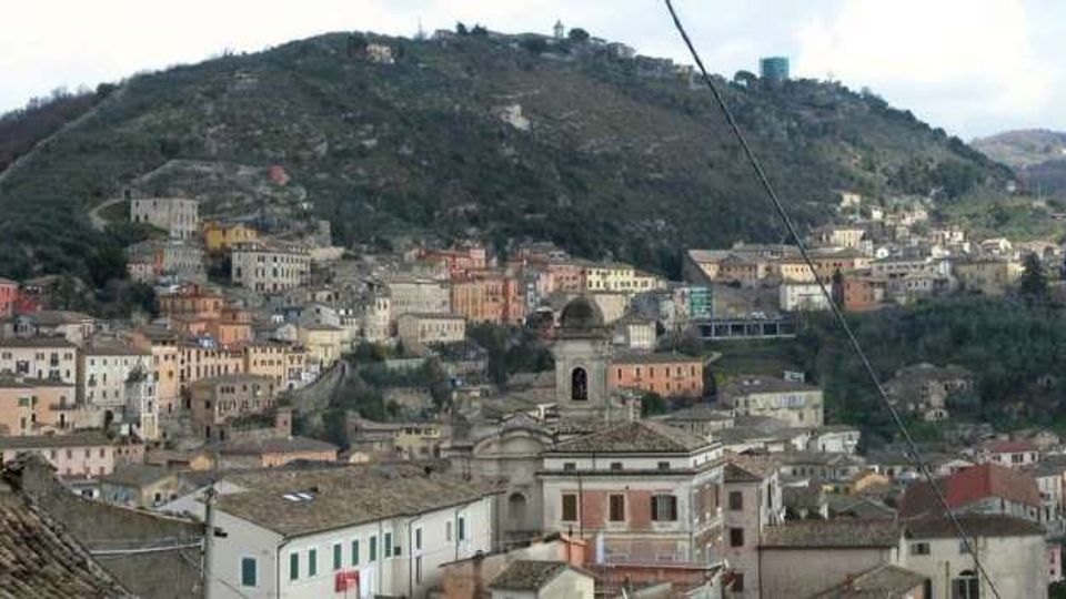 Panorama di un borgo collinare con case addossate su un pendio, tetti in coppi e un campanile in primo piano. Sullo sfondo si vede una grande collina boscosa con strade serpeggianti e alcune strutture sulla cresta.
