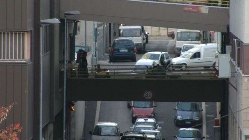 Strada urbana stretta tra edifici alti, con traffico intenso di auto e furgoni. Sopra la carreggiata si vedono due passaggi pedonali sopraelevati che collegano gli edifici, con corrimano e strutture in metallo.