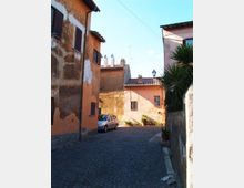 Vicolo stretto in pietra con pavimentazione in sampietrini, fiancheggiato da edifici intonacati color ocra e rosa con persiane scure e tetti in coppi. In fondo si vedono alcune piante in vaso, un lampione e una piccola auto parcheggiata lungo il muro.