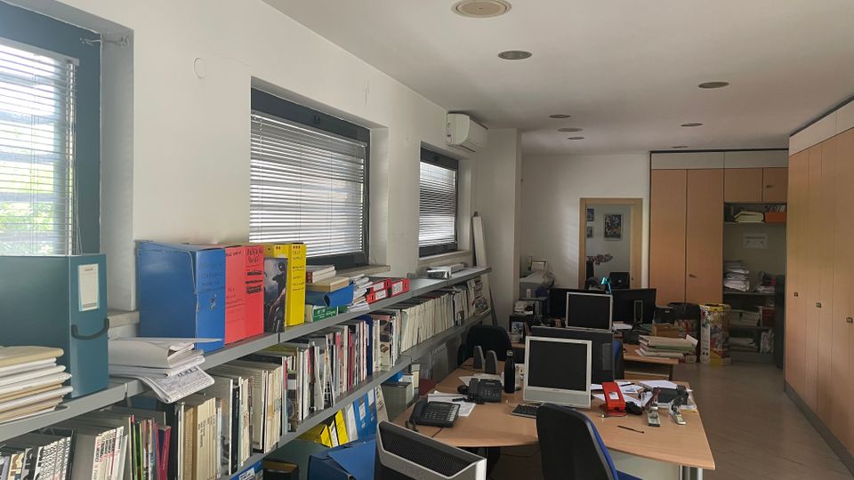 Interno di un ufficio allungato con scaffalature piene di libri e raccoglitori lungo il lato sinistro sotto finestre con veneziane. Sulla destra ci sono postazioni con scrivanie, computer e sedie, armadi alti in legno e illuminazione a faretti incassati nel soffitto.