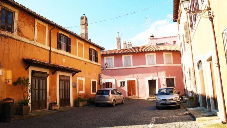 Piccola piazza acciottolata in un borgo, circondata da edifici bassi dai colori caldi (arancione e rosa) con persiane, porte in legno e tetti in tegole. Sono presenti lampioni a parete, alcuni vasi di piante e auto parcheggiate lungo i lati.