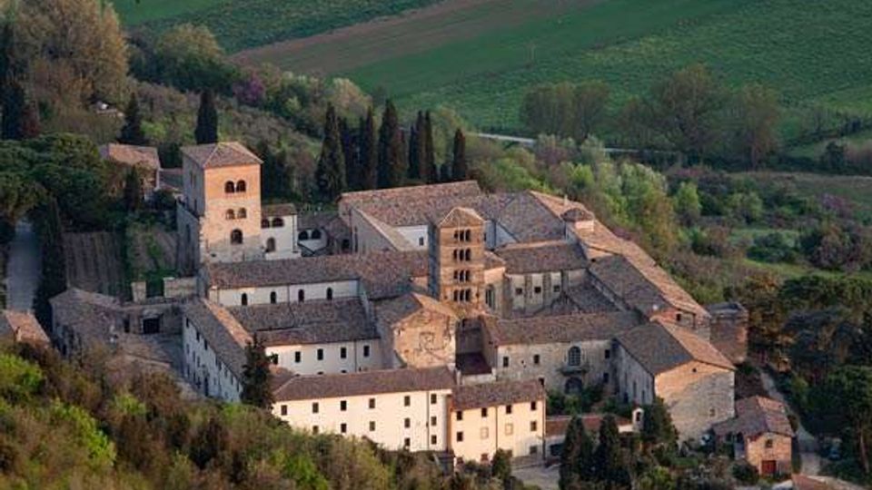Veduta aerea di un complesso monastico in pietra con vari edifici a tetti in tegole e un campanile centrale, disposto attorno a cortili interni. Il monastero è circondato da alberi e colline con campi coltivati sullo sfondo.