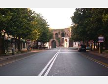 Strada urbana a due corsie con doppia linea continua centrale, fiancheggiata da alberi e edifici bassi. In fondo si vede una grande struttura in mattoni con archi e aperture, simile a una porta o resti di fortificazione.