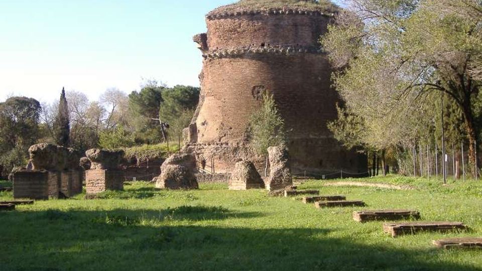 Grande torre circolare in mattoni antichi, con sommità coperta di vegetazione, in un’area archeologica erbosa. In primo piano si vedono resti di muri e blocchi di pietra sparsi, con alberi e recinzioni sullo sfondo.