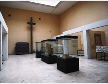 Sala museale con pareti beige e soffitto alto, illuminata da un lucernario, con tre teche in vetro su basi nere al centro. Sul fondo si vede una grande croce nera e un grande sarcofago scolpito, con altre opere esposte su piedistalli lungo le pareti.