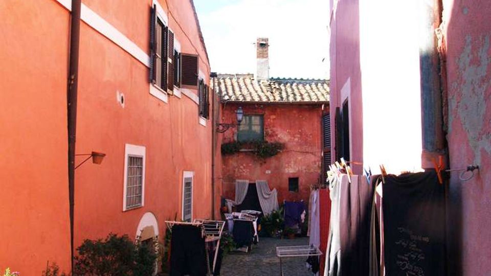 Vicolo stretto con pavimentazione in ciottoli tra edifici intonacati color arancio e rosa, con tetti in coppi e finestre con persiane. Lungo i muri si vedono vasi con piante e stendibiancheria con panni appesi, con vista su una piccola corte in fondo.