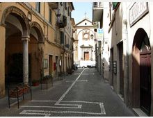 Via stretta in un centro storico con pavimentazione in pietra, edifici alti ai lati e portici ad archi con colonne sul lato sinistro. In fondo alla strada si vede la facciata di una chiesa con timpano e rosone, con un veicolo parcheggiato davanti.
