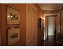 Corridoio interno di una casa con pareti color ocra, pavimento scuro e porte bianche a pannelli lungo il passaggio. Alle pareti sono appesi quadri incorniciati e una lampada a parete illumina l’ingresso verso una porta in fondo.