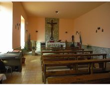 Interno di una piccola cappella con pareti color pesca e rivestimento in pietra nella zona dell’altare, con crocifisso centrale sopra l’altare. File di panche in legno su pavimento piastrellato, luce laterale da una finestra e alcune decorazioni religiose alle pareti.
