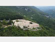 Veduta aerea di un grande complesso monastico in pietra con tetti in laterizio, disposto attorno a cortili e racchiuso da mura. L’edificio sorge su un pendio montano ed è circondato da fitti boschi, con vallate e montagne sullo sfondo.