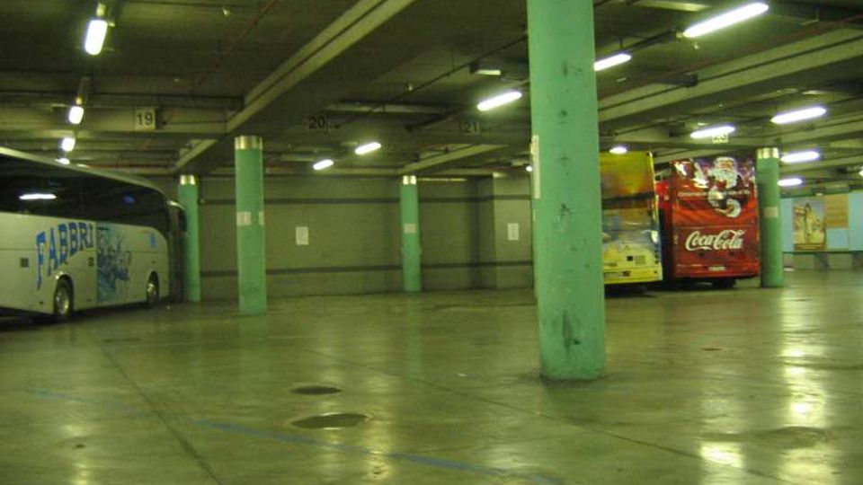 Ampio parcheggio sotterraneo o deposito per autobus con pavimento in cemento lucido, colonne cilindriche verde chiaro e illuminazione a neon sul soffitto. Alcuni autobus sono parcheggiati ai lati, con numeri di corsia visibili sulle travi.