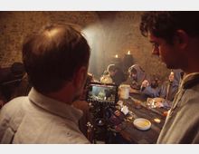 Interno in pietra di una taverna o cantina rustica, con volta in muratura e un lungo tavolo di legno illuminato da candele. In primo piano si vede una videocamera con monitor che inquadra la scena del tavolo con stoviglie e brocche.