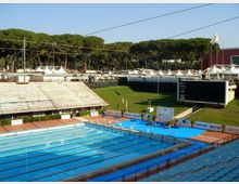 Stadio del nuoto all’aperto con piscina olimpionica a corsie, tribune in cemento e piattaforme per tuffi sul bordo. Sullo sfondo si vedono un grande tabellone elettronico, un pendio erboso, cavi di copertura e una fila di alberi alti oltre le strutture.