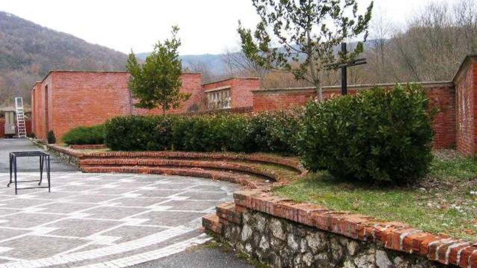 Cortile esterno pavimentato con disegni geometrici, delimitato da muretti in pietra e mattoni e aiuole con siepi e alcuni alberi. Sullo sfondo si vedono edifici bassi in mattoni rossi e colline boscose.