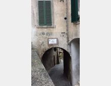Stretto vicolo in pietra che scende sotto un arco ricavato tra edifici antichi con intonaco consumato e crepe. Sulla facciata si vedono persiane verdi e un cartello stradale “Via Epifania”, con cavi esterni e una piccola apertura circolare nel muro.