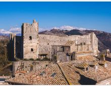 Castello medievale in pietra su un’altura, con alte mura e una torre quadrata. In primo piano si vedono tetti di case con coppi in terracotta, sullo sfondo colline e montagne sotto cielo sereno.