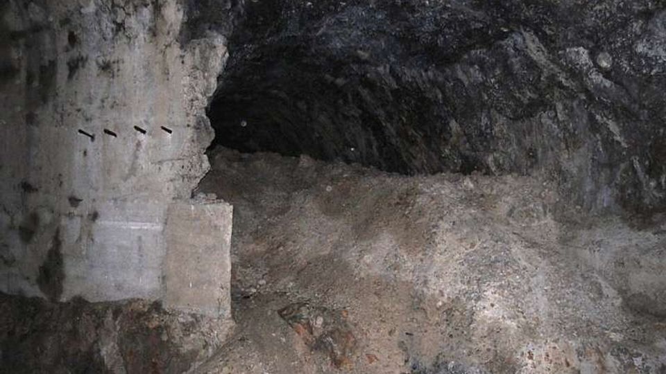 Interno di una galleria sotterranea in roccia grezza, con pareti irregolari e annerite e un piccolo passaggio ad arco che prosegue nel buio. Il pavimento è coperto da cumuli di terra e detriti, con un tratto di muratura o intonaco sul lato sinistro.