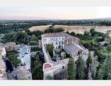 Veduta aerea di un complesso fortificato in pietra con cortile interno alberato e tetti in coppi, circondato da mura con torri. Intorno si vedono un parcheggio e edifici del borgo, oltre a campi coltivati e boschi nella campagna circostante.