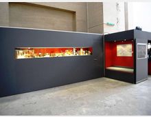 Allestimento museale in uno spazio interno con pareti grigio scuro e una lunga vetrina orizzontale illuminata che espone oggetti. A destra c’è una nicchia con pareti rosse, panca integrata e un pannello espositivo, su pavimento in cemento.